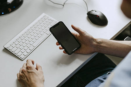 Jemand am Schreibtisch mit Tastatur, hält ein Smartphone in der Hand.