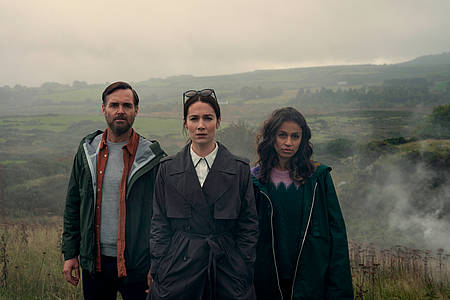 Die drei True Crime Podcaster stehen im nebeligen Moor und starren in die Ferne - Bodkin auf Netflix. 