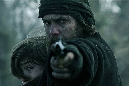 Ein bärtiger Mann drückt ein Kind an sich und richtet seinen Pistolenlauf auf die Kamera - American Primeval auf Netflix.